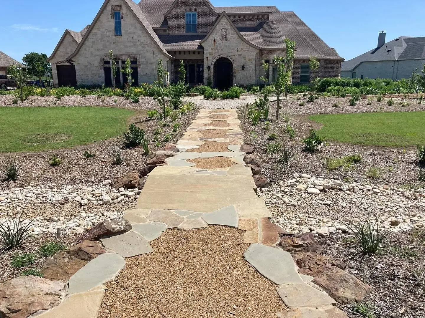 A decomposed granite walkway edged with flagstone and boulders leading to a stone-and-brick home, flanked by newly planted perennial beds and river rock