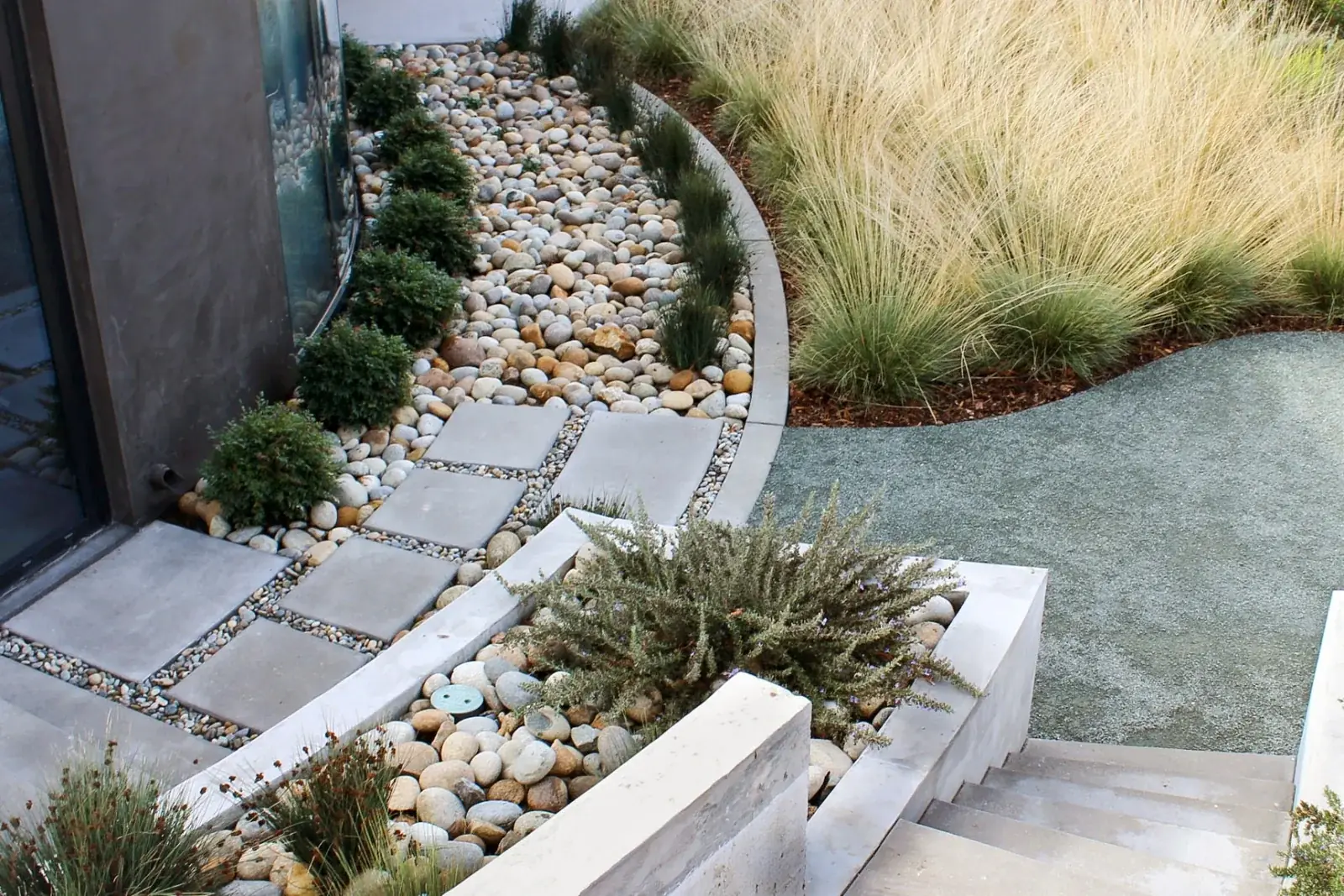 Concrete stepping pads set in graded river rock beside a modern home, with board-formed concrete edging and drifts of ornamental grass