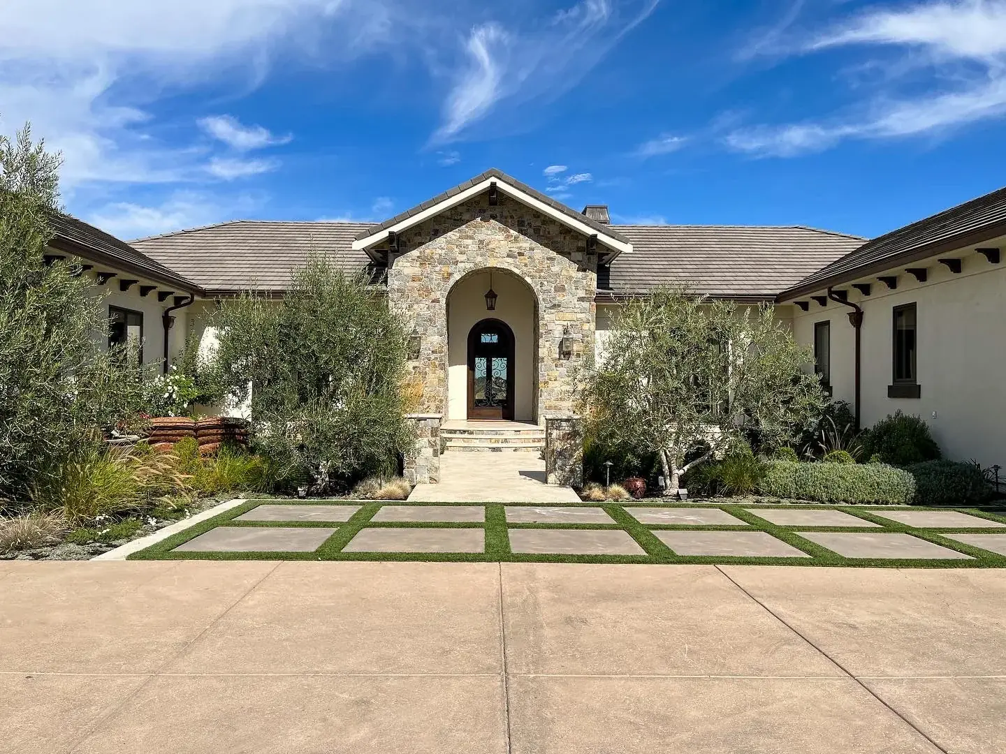 Oversized concrete pavers set in strips of turf leading from a driveway to a stone-arched entry, lined with multi-trunk olive trees and silver-toned planting
