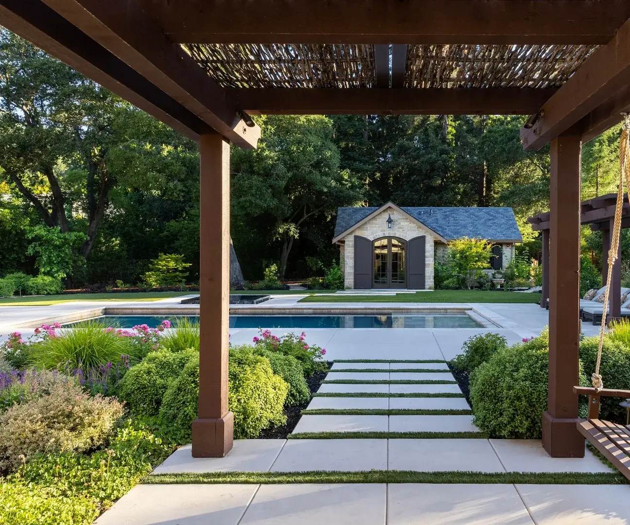 A pool terrace seen from under a cedar shade structure, with large-format pavers set in planted joints and layered evergreen and perennial beds around the water